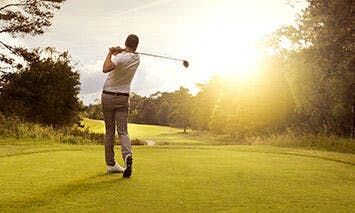 A golfer swings a club on a well-maintained course, surrounded by trees and greenery, with the sun shining brightly in the background.